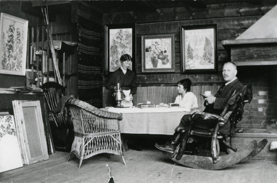 Maija Halonen, rouva Nybom ja Pekka Halonen teehetkellä Halosenniemen ateljeen takkanurkassa vuonna 1915. Kuvan lisenssi: CC BY 4.0.
